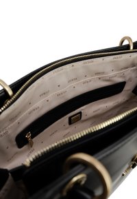 Borsa nera in pelle con zip superiore. L'interno presenta una fodera beige con il logo "GUESS" ripetuto e varie tasche, tra cui una tasca con zip.