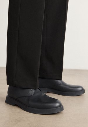 Chaussures à lacets en cuir noir portées avec un pantalon noir à jambes larges, debout sur un sol lisse marron clair.