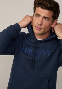 Marineblauwe hoodie met trekkoord, voorzien van een voorzak en blauwe "HACKETT LONDON" branding. Zachte stof met een klassieke snit.