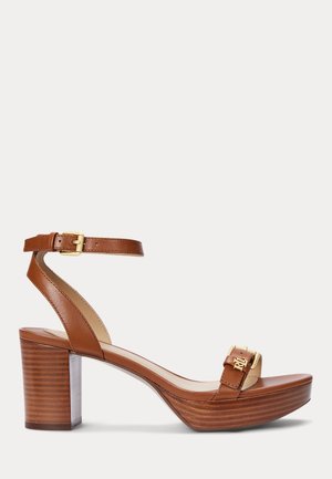 Brun læder højhælet sandal med ankelrem, guldbeslag, platformsål og blokhæl på en hvid baggrund.