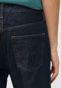 Άτομο που φοράει σκούρα μπλε τζιν με ορατές πορτοκαλί ραφές και τυρκουάζ πράσινο πουκάμισο μέσα από το παντελόνι.