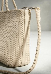 Sac à main en cuir beige tissé avec une bandoulière tressée et une fermeture argentée sur une surface et un fond gris clair.