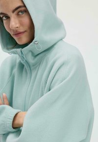 Hellblaues Fleecepullover mit hohem Kragen, großer Kapuze und Reißverschluss. Verfügt über weite Ärmel und eine weiche, strukturierte Oberfläche.