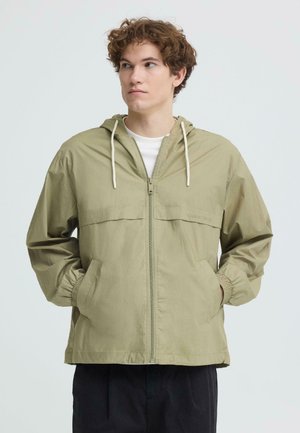 Junger Mann mit lockigem Haar, der eine hellgrüne Kapuzenjacke mit Reißverschluss trägt und die Hände in den Taschen hat, blickt auf einem einfarbigen Hintergrund nach links.