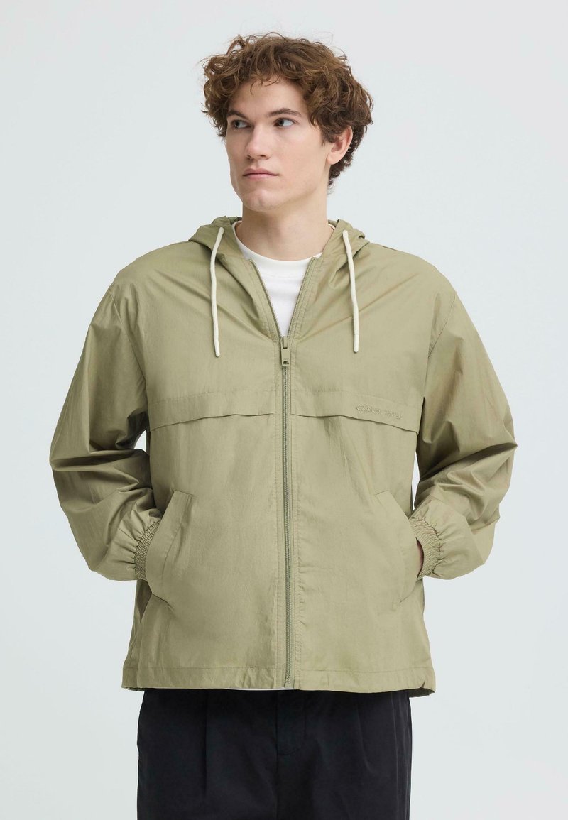 Młody mężczyzna o kręconych włosach, ubrany w jasnozieloną kurtkę z kapturem zapinaną na zamek, z rękami w kieszeniach, patrzący w lewo na jednolitym tle.