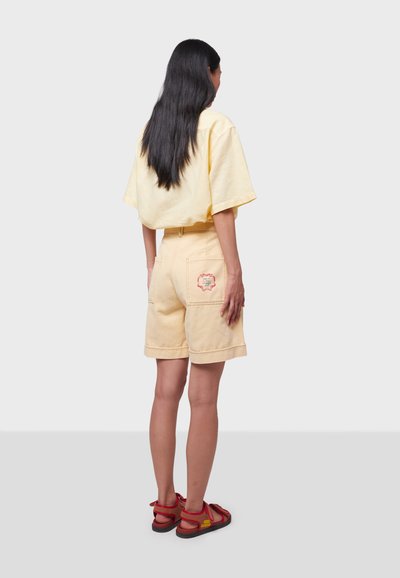 T-shirt oversize en coton jaune associé à un short assorti, doté de deux poches arrière et d'un détail brodé rouge. Des sandales marron complètent le look.