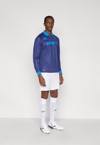 Maglia da calcio a maniche lunghe color blu navy con strisce verticali, colletto blu e stampa a contrasto. Abbinata a pantaloncini e calzini bianchi, scarpe da calcio nere.