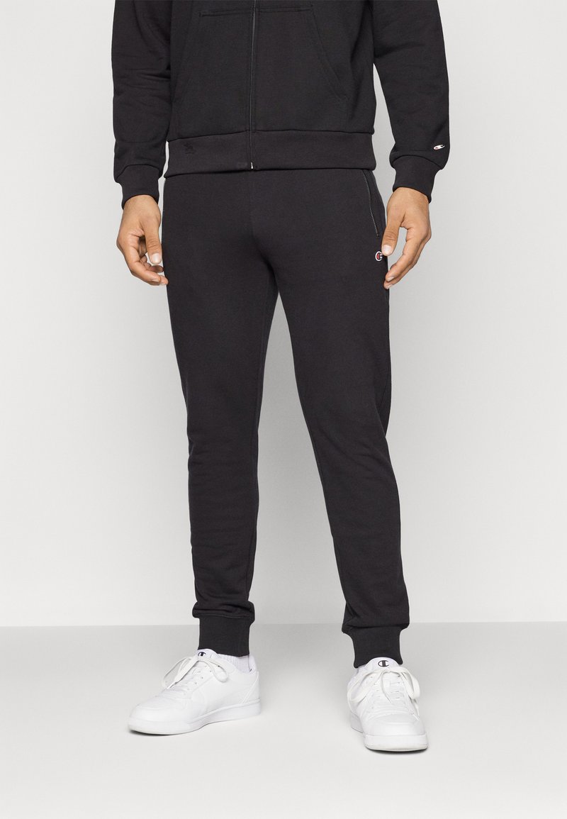 Pantalons de survêtement noirs avec un design ajusté, taille élastique et poignets, associés à des chaussures de sport blanches. Présente un petit logo sur le côté.