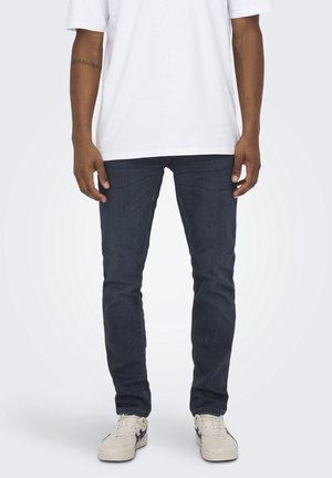 Person trägt dunkelblaue Slim-Fit-Jeans, weiße Sneakers und ein schlichtes weißes kurzärmeliges T-Shirt und steht vor einem neutralen Hintergrund.