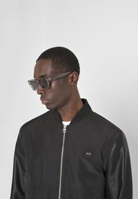 Black bomber jacket with a zipper, featuring fabric sleeves, a logo patch on the chest, and worn with gray rectangular sunglasses.