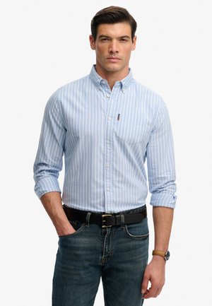 Mann trägt ein hellblau gestreiftes Hemd mit hochgekrempelten Ärmeln, dunkle Jeans, schwarzen Gürtel und braune Uhr, steht mit einer Hand in der Tasche.