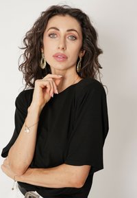 Czarny krótki top z luźnym krojem, gładka faktura. Złote wyraziste kolczyki, warstwowe bransoletki z okrągłymi zawieszkami i naturalny makijaż.