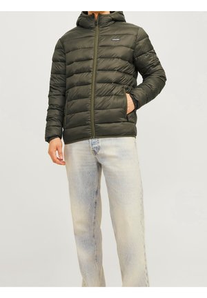 Mann trägt eine olivgrüne gesteppte Jacke mit Reißverschluss und Kapuze sowie hellblaue, ausgewaschene Jeans, Hand in der Jackentasche.