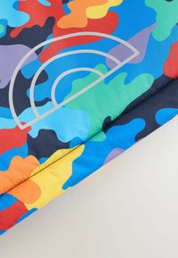 Farbige Tarnjacke mit auffälligen Spritzern in Rot, Blau, Grün und Gelb, versehen mit einem hellgrauen runden Logo auf einem dunklen Hintergrund.
