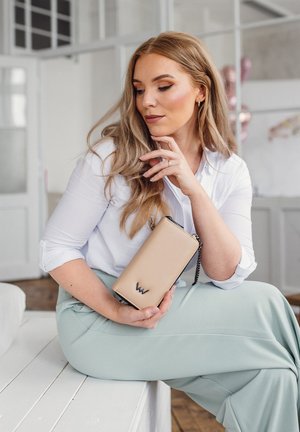 Beige wallet with a black logo, made of textured material. It features a chain strap and is held against a light-colored outfit.