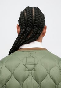 Jachetă matlasată verde cu guler rotunjit. Prezintă un model texturat în valuri și cusături vizibile, completat de un guler și detalii de culoare maro.