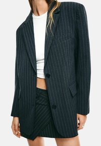 Donna con indosso un blazer a righe scure e una gonna abbinata sopra una maglietta bianca corta, che mostra il ventre e indossa un anello d'argento.