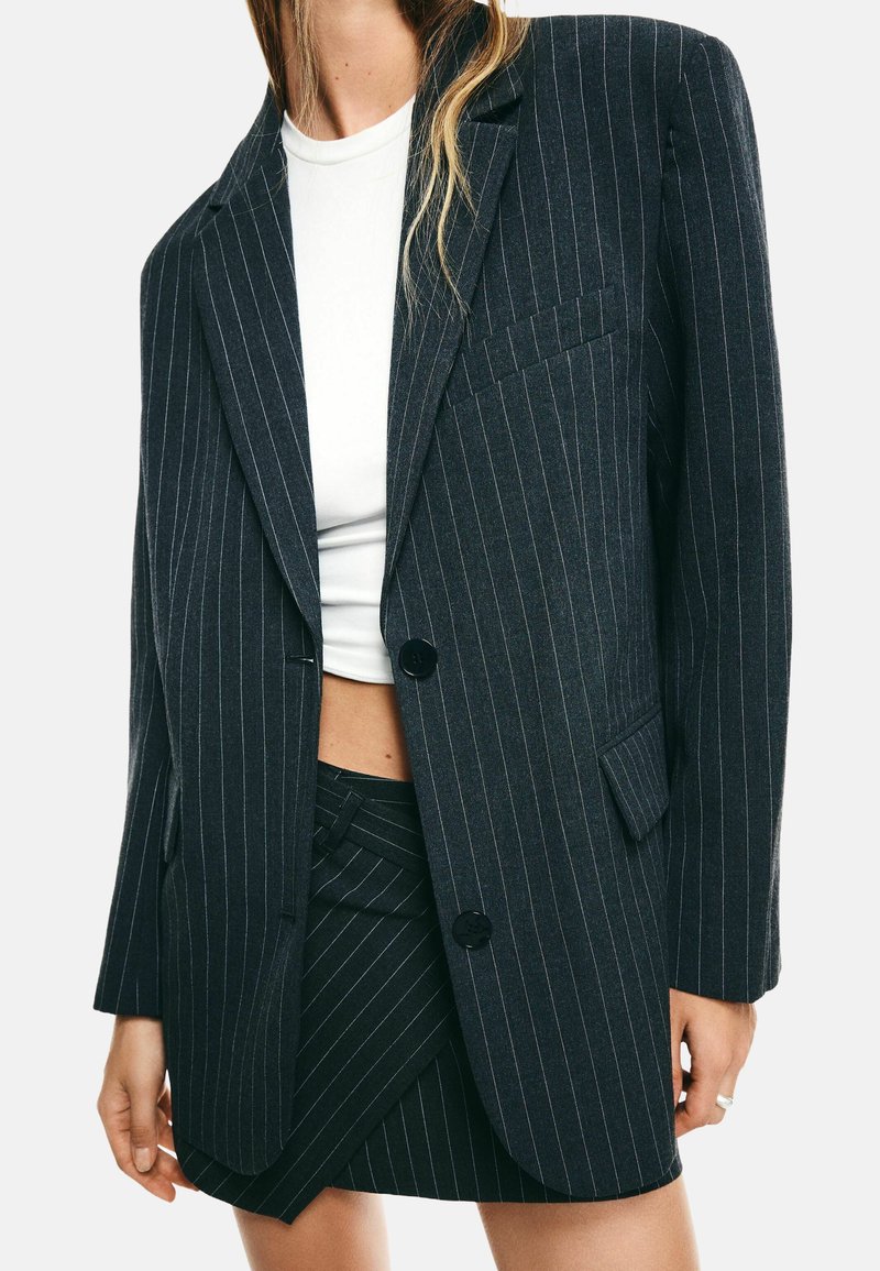 Donna con indosso un blazer a righe scure e una gonna abbinata sopra una maglietta bianca corta, che mostra il ventre e indossa un anello d'argento.
