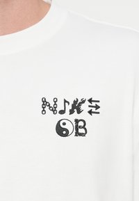 Λευκό βαμβακερό t-shirt με μαύρα κεντημένα γραφικά στοιχεία: αλυσίδα, μουσική νότα, φλόγες, βέλη και σύμβολο γιν γιανγκ.