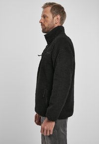 Giacca in pile nera con colletto alto; presenta una tasca con zip e due tasche laterali. Tessuto testurizzato, vestibilità rilassata e maniche lunghe.