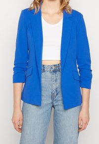 Blå blazer med långa ärmar, smala slag och två framfickor. Buren över en vit crop top och parat med högmidjade denimjeans.