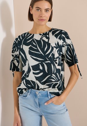 Femme portant un haut blanc à manches courtes avec de grands motifs de feuilles noires et un jean bleu clair, debout avec une main dans la poche.