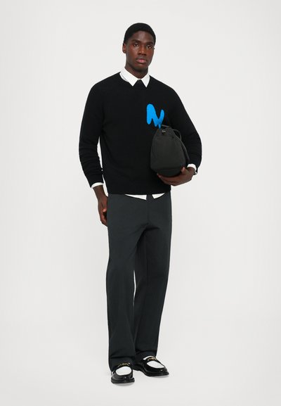 T�ânăr îmbrăcat cu un pulover negru cu un "N" albastru, ținând o geantă neagră, cămașă cu guler alb, pantaloni negri și mocasini alb-negru cu lanț de aur.