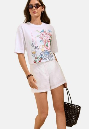 Nainen, jolla on valkoiset shortsit ja valkoinen t-paita, jossa on värikäs kirjailu kukista, kynttilöistä, lautasella olevasta kalasta, ja hän pitää mustaa kudottua laukku.