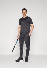 Zwart sportoutfit met een geblokte T-shirt en broek, gemaakt van lichtgewicht stof. Inclusief een tennisracket en zwarte sneakers.