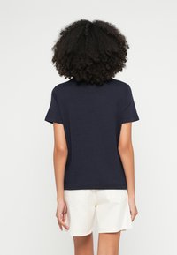 Tmavomodré bavlnené tričko s krátkymi rukávmi, okrúhlym výstrihom a voľným strihom, v kombinácii s krémovobéžovými šortkami. Hladká textúra, minimalistický dizajn.