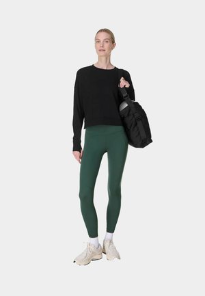 Femme en haut à manches longues noir, leggings verts, baskets blanches, portant un sac de sport noir avec un casque à l’intérieur, debout sur fond blanc.