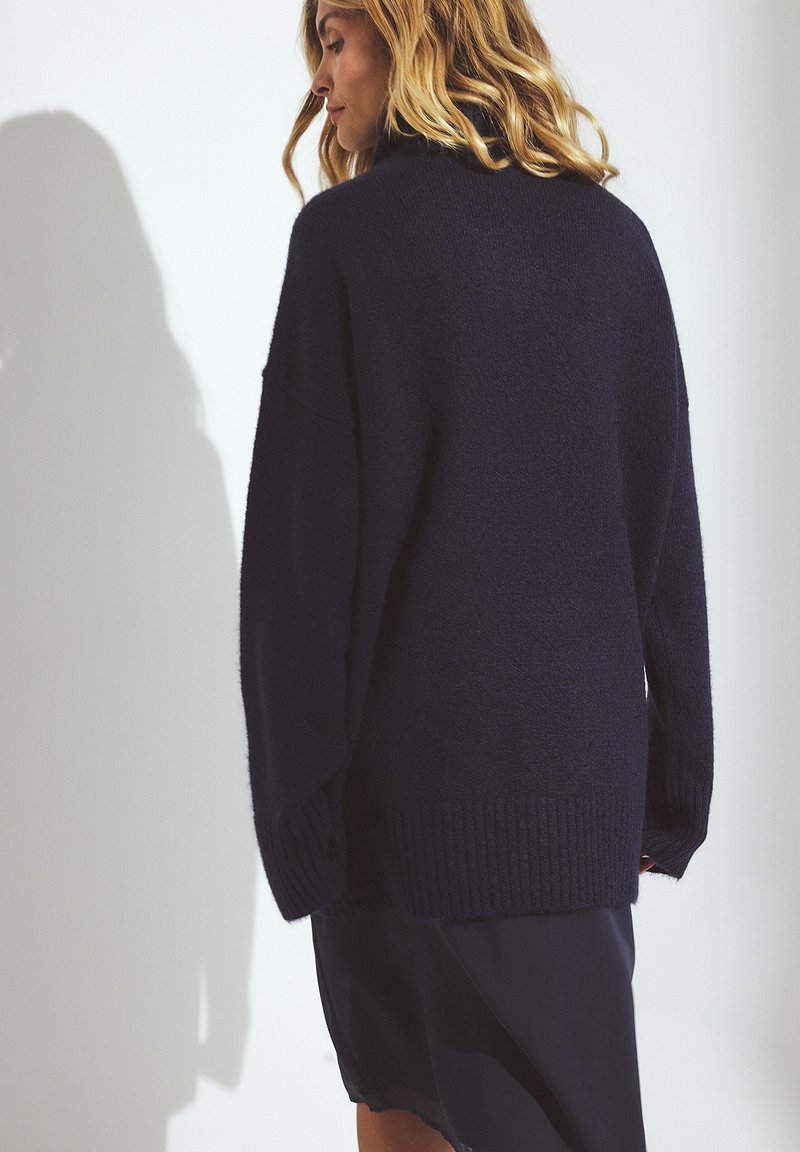 Pull en maille bleu marine surdimensionné en matière douce et fluffy, avec des bords côtes aux poignets et à l'ourlet. Conçu avec un col montant et des épaules tombantes pour une coupe décontractée.
