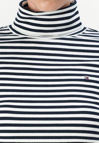 Top cu guler înalt, cu dungi navy și albe, textură canalată, cu un mic logo brodat pe pieptul stâng. Design ajustat.