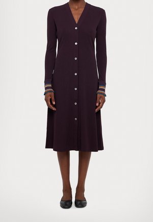 Donkerpaarse jurk met lange mouwen, knopen aan de voorkant, V-hals en kleurrijke gestreepte manchetten, gedragen met zwarte platte schoenen.