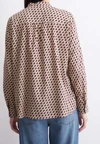 Bluzka z długim rękawem wykonana z materiału w wzór, z małymi bordowymi diamentami na beżowym tle, z prostym dołem i mankietami zapinanymi na guziki.