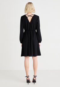 Μαύρο φόρεμα με μακριά μανίκια, ελαστική μέση και ανοιχτή πλάτη με σταυρωτές τιράντες. Λείος ύφασμα και μήκος μέχρι το γόνατο. Φορεμένο με μαύρες γόβες.