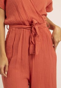 Jumpsuit texturé de couleur rouge framboise avec un design enroulé, des manches courtes et une ceinture en tissu. Présente des détails plissés à la taille.