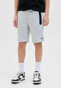 Pantaloni corti in cotone grigio chiaro con vita elastica, tasche laterali con zip e un piccolo logo nero sul lato sinistro in basso. Indossati con scarpe da ginnastica nere.