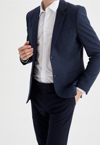 Blazer navy strutturato con rever inclinati, chiusura a un bottone e silhouette aderente, abbinato a una camicia bianca con bottoni e pantaloni scuri.