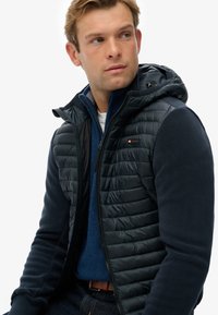 Marineblauwe hybride jas met een gewatteerd bovenstuk, fleece mouwen en een ritsluiting met een capuchon. Gedetailleerde stiksels zichtbaar.