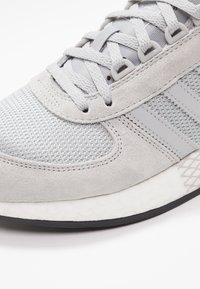 Sneaker grigio con tomaia in rete e dettagli in suede, lace tonali e una suola intermedia bianca testurizzata. Suola in gomma nera visibile.
