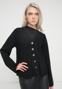 Cardigan negru tricotat cu decolteu în V, dotat cu cinci butoane metalice și pliuri verticale pe față. Mâneci lungi, finisaj texturat.