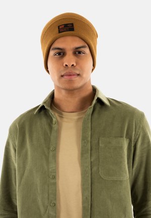 Bonnet marron à côtes avec une étiquette noire et orange ; chemise en velours côtelé olive avec des boutons sur le devant et une poche plaquée sur un t-shirt beige.