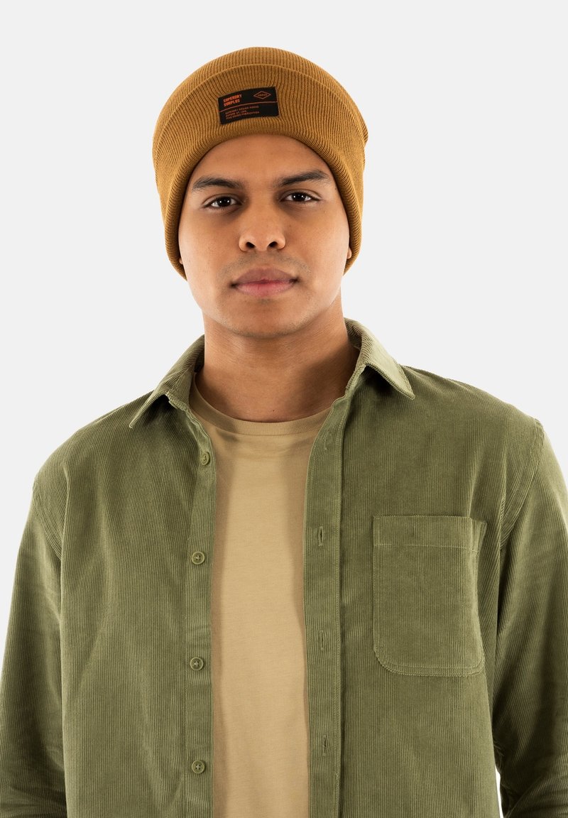Gorro de punto marrón con etiqueta negra y naranja; camisa de pana verde olivo con botones y un bolsillo de parche sobre una camiseta beige.