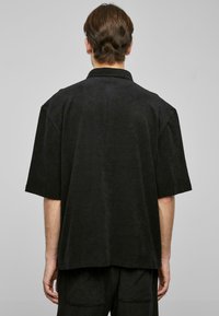 Hombre con cabello corto castaño, vistiendo una camisa negra de manga corta holgada y pantalones negros a juego, de espaldas frente a un fondo gris liso.