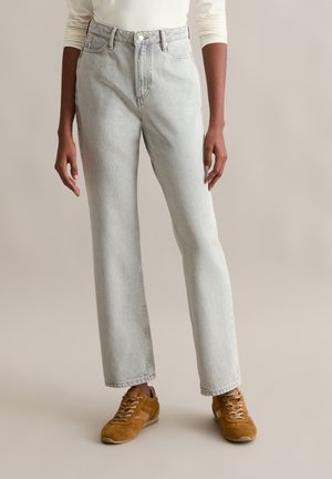 Jean taille haute, délavé clair, avec une coupe droite, comportant cinq poches et une fermeture à bouton. Porté avec des baskets en daim marron.