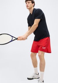 Μαύρο αθλητικό μπλουζάκι με κοντά μανίκια, κόκκινα σορτς με λογότυπο, λευκές κάλτσες και παπούτσια τένις. Κρατάει ρακέτα τένις, έτοιμος για παιχνίδι.