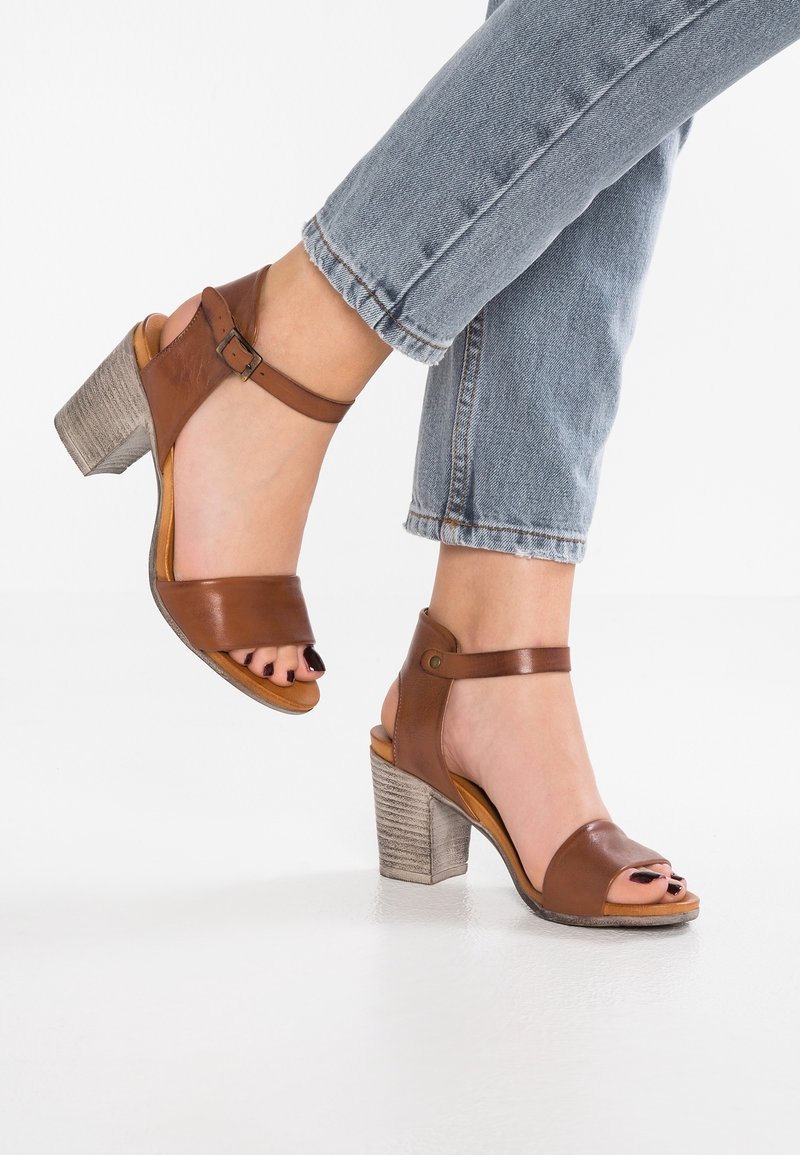 Personne portant des sandales à talons carrés en cuir marron avec des brides à la cheville et un jean en denim bleu raccourci, sur un fond de sol blanc.