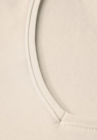 Světle béžová textilie s hladkým povrchem, s širokým kulatým výstřihem a viditelnými stehy podél okrajů.