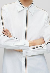 Bílá košile s klasickým límcem, zdobená černými a hněd ými vertikálními pruhy podél zapínání a manžet. Tlačítka jsou bílá a kulatá.